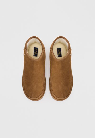 Papuci din suede maro, cu o căptușeală interioară moale, design rotunjit și detalii minime de brăzdare. Prezintă un mic logo pe o parte.