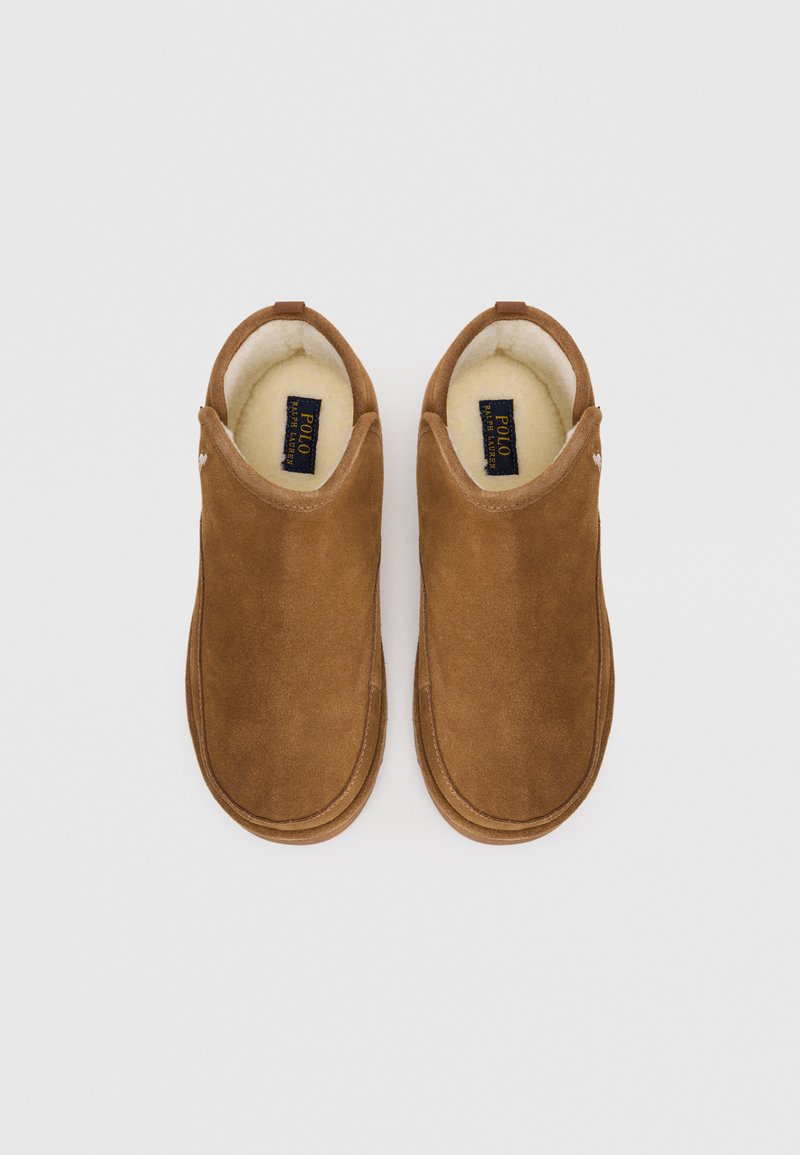 Chaussons en suede marron avec un intérieur doux, un design arrondi et des détails de couture minimalistes. Présente un petit logo sur un côté.