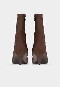 Καφέ μποτάκια από σουέντ με καμπυλωτό τακούνι, λείο υφή και δεμένα διακοσμητικά στο πάνω μέρος, φαίνονται από πίσω.