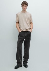 Maglietta beige a maniche corte abbinata a jeans grigio scuro a gamba dritta; il modello è in piedi con le mani nelle tasche, indossa scarpe nere.