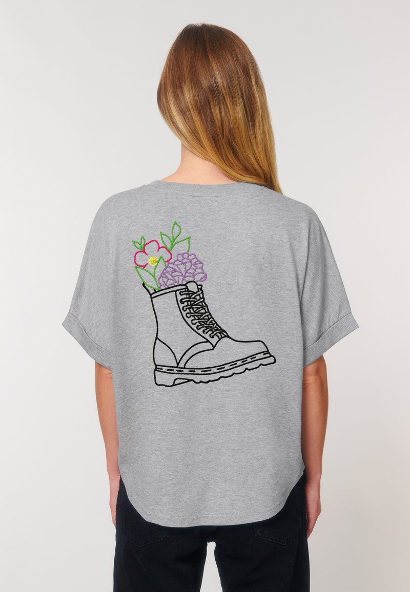 Chandail gris à manches courtes avec un contour noir d'une botte, orn é de fleurs colorées émergeant de celle-ci au dos. Coupe décontractée et texture douce.