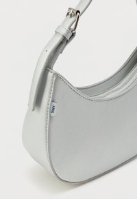 Sudraba sintētiska plecu soma ar izliekta dizainu, spīdīgu apdari un rāvējslēdzēju. Ievērojama maza balta birka ar zilu tekstu.