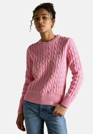 Frau trägt einen pinkfarbenen Kabelstrickpullover und blaue Jeans, posiert mit einer Hand in der Tasche vor einem einfarbigen hellen Hintergrund.