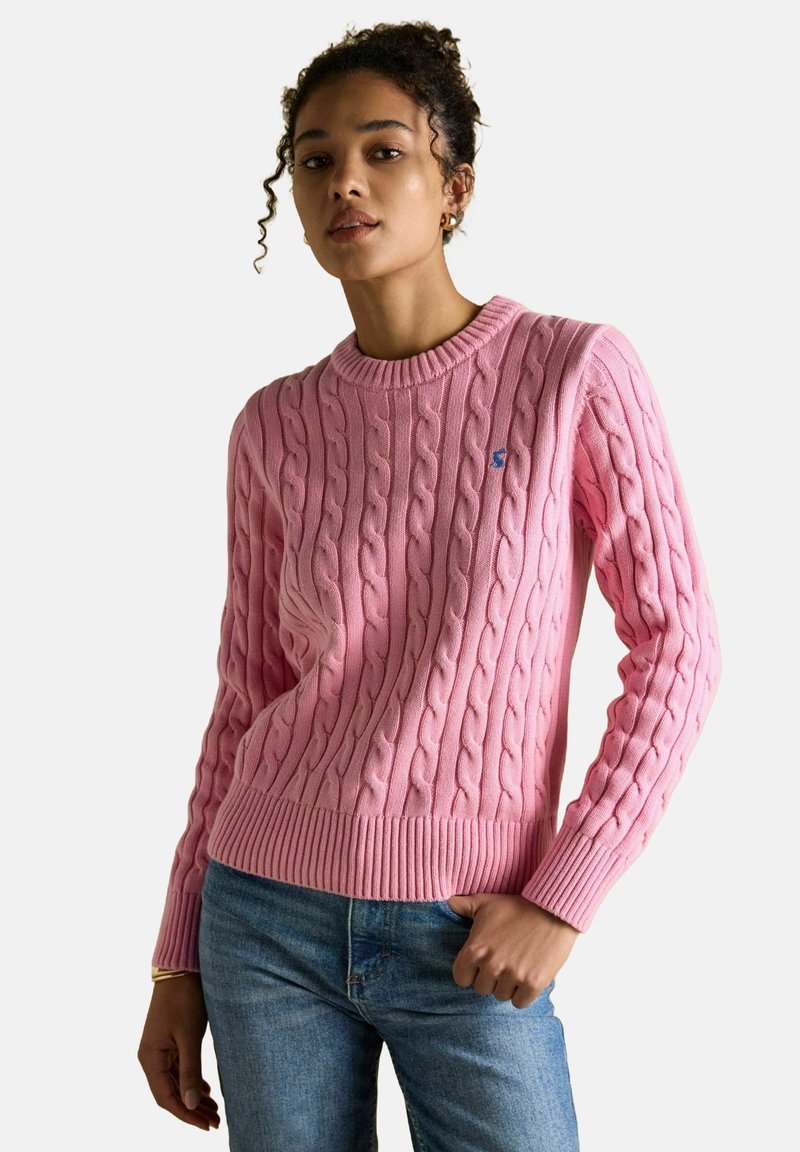 Kobieta w różowym swetrze w warkocze i niebieskich dżinsach, pozująca z jedną ręką w kieszeni na jednolitym jasnym tle.