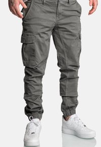 Mężczyzna w szarych spodniach dresowych cargo i białych trampkach Nike, stojący z rękami w pobliżu kieszeni na białym tle.