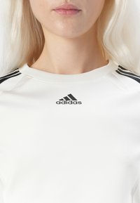 Weißes Sporttop mit schwarzen Drei-Streifen-Akzenten an den Schultern und dem Adidas-Logo in Schwarz. Glatte Textur mit rundem Halsauschnitt.