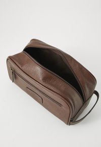 Estojo de toalete de couro marrom com um zíper na parte superior aberto, bolso frontal com zíper e alça lateral para transporte, sobre um fundo branco.