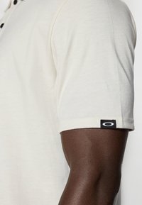 Polo shirt beige clair en tissu doux, doté d'un col standard et d'une étiquette logo noire sur la manche. Vue rapprochée de la manche et du bras.