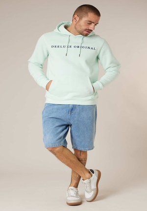 Homme portant un sweat à capuche vert menthe clair avec l'inscription "DEELUXE ORIGINAL", un short en jean et des baskets blanches, debout les mains dans les poches et les jambes croisées.