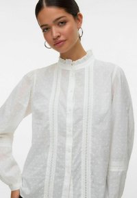 Witte katoenen shirt met een gerimpelde kraag, knoopsluiting en een textuur van stippen. De mouwen zijn licht geblokt met details.