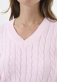 Heled roosa soonkootud sviiter V-kaelusega, millel on tekstuursed mustrid ja ribakraed. Materjal näeb välja pehme ja kerge.