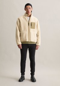 Jonge man in een beige fleecejack met ritszakken, zwarte jeans, zwarte laarzen en een achterwaartse zwarte pet, staand tegen een effen achtergrond.