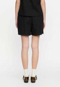 Svarta shorts i ögla tyg, scalloped nederkant och lös passform. Bärs med bruna skor och vita strumpor. Enkelt design, utan mönster eller detaljer.