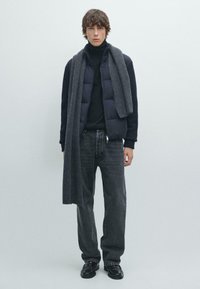 Mladý muž stojící v tmavé prošívané bundě, černém roláku, tmavě šedých džínách, černých mokasínech a dlouhém šedém pleteném šátku.