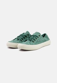 Zelene platnene tenisice s zaobljenim nosovima, teksturiranom površinom, bijelim gumenim potplatima i metalnim rupicama za vezice. Minimalistički dizajn.