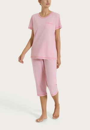 Donna in piedi scalza che indossa un pigiama composto da top a maniche corte rosa chiaro e pantaloni corti con rifiniture rosa più scuro e una piccola tasca sul petto.