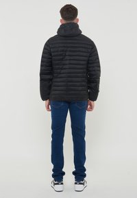 Giacca di piumino nera con cappuccio, con texture trapuntata. Indossata sopra jeans blu e sneakers nere, vista da dietro.