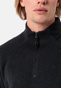 Črn zip-up jumper z rebrasto strukturo, visokim ovratnikom in kratkim zadrgo. Ima subtilno blagovno znamko na levi strani.