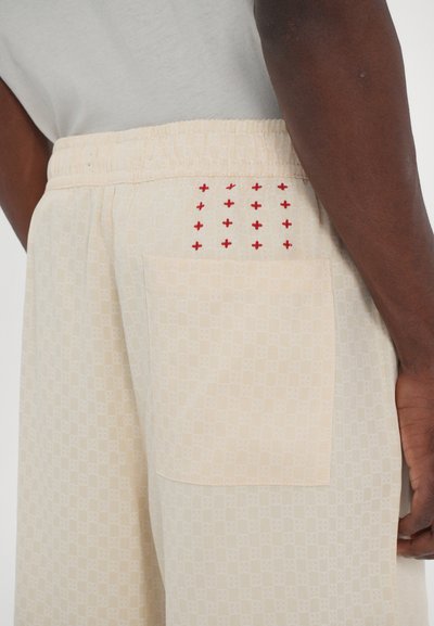Lichtbeige sweatpants met een gestructureerd patroon, voorzien van een achterzak met rode kruissteekaccenten en een elastische tailleband.