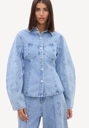 Femme portant une veste en jean bleu clair avec des coutures décoratives sur les manches et un jean taille haute assorti sur fond blanc.