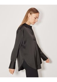 Donna di profilo che indossa una blusa nera in satin morbido con polsini oversize e un orlo curvo, in piedi su uno sfondo bianco semplice.