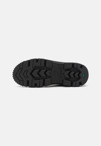 Semelle de botte en caoutchouc noir avec un motif de profondeur, présentant un logo central et une petite touche de vert. Surface texturée pour une meilleure adhérence.