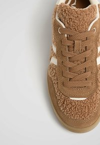 Baskets marron en tissu duveteux texturé, détails en daim, accents crème et lacets tissés sur un fond clair.