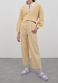 Frau, die eine cremefarbene Häkeljacke, karierte hochtaillierte weite Hose, lilafarbene Socken und lilafarbene Sandalen mit offenen Zehen trägt, steht auf einem grauen Boden.