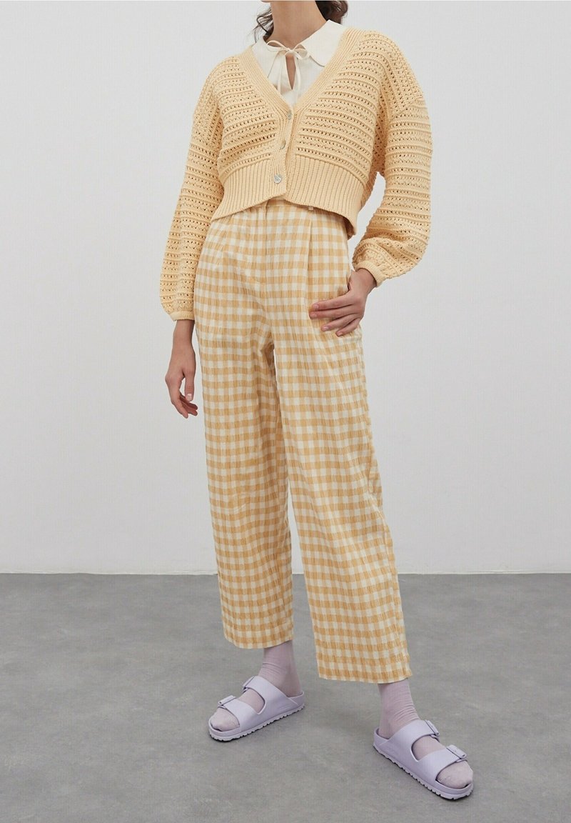 Frau, die eine cremefarbene Häkeljacke, karierte hochtaillierte weite Hose, lilafarbene Socken und lilafarbene Sandalen mit offenen Zehen trägt, steht auf einem grauen Boden.