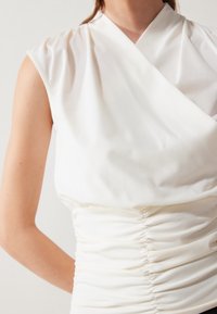Blusa blanca sin mangas con cintura fruncida y amplio cuello envolvente, llevada por una persona con brazo desnudo y tono de piel claro.