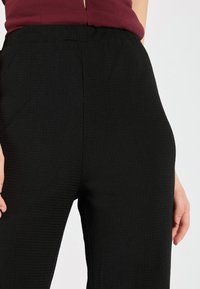 Black textured trousers with a relaxed fit. The fabric features a subtle pattern. Elastic waistband visible, paired with a maroon top.
