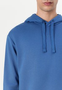 Modra kapuca iz mehkega blaga, z vrvico za prilagajanje na kapuci in diskretnim logotipom. Gladka tekstura z udobnim krojem in dolgimi rokavi.