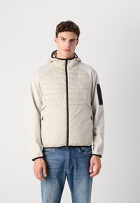 Lightweight, beige puffer jacket with ripstop texture, hood, front zip, and black accents. Features side pockets and elastic cuffs.