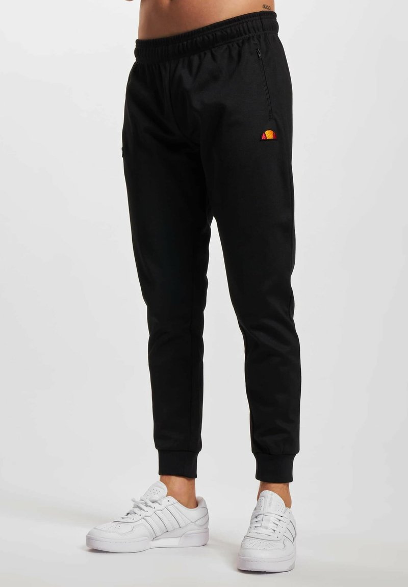 Pantalones de chándal negros con una cinturilla elástica, corte ajustado y bolsillos laterales. Presenta un pequeño logotipo multicolor en el muslo izquierdo. Se llevan con zapatillas blancas.