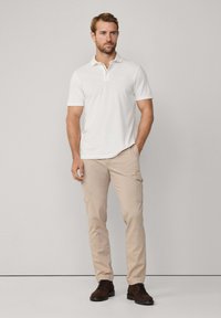 Polo shirt bianco con colletto, abbinato a pantaloni cargo beige con tasche laterali. Scarpe marroni con lacci completano l'outfit.