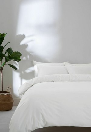 Weißes Bett mit Kissen und Bettdecke neben einer grünen Topfpflanze in einem geflochtenen Korb, deren Schatten auf eine schlichte weiße Wand fällt.