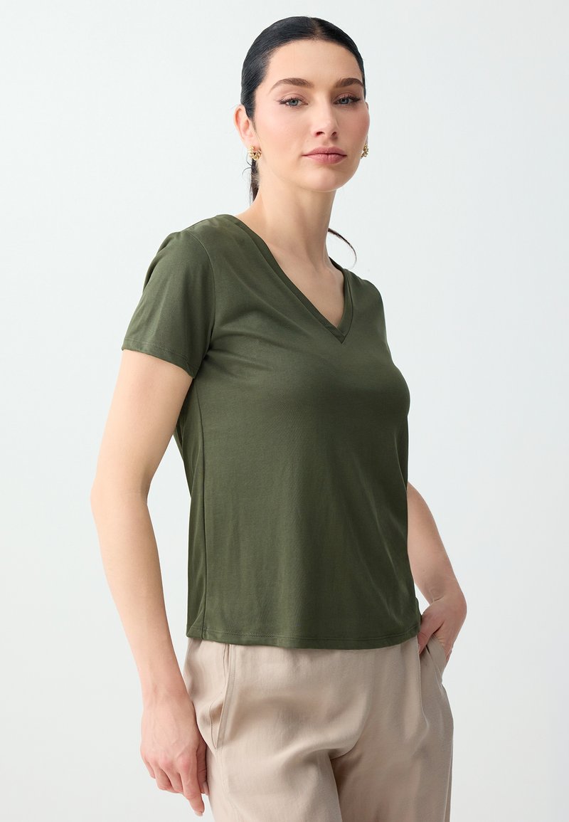Femme aux cheveux foncés portant un t-shirt vert à col en V et manches courtes et un pantalon beige, debout avec une main dans la poche devant un fond clair uni.