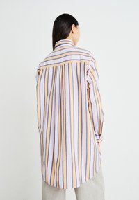 Chemise longue rayée en rose, jaune et bleu. Présente un col boutonnière, des manches longues, un pli au dos et un ourlet arrondi. Fabriquée en tissu léger.