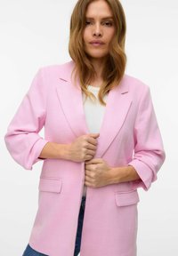 Blazer de color rosa claro con tejido texturizado, cierre de un botón, bolsillos con solapa y mangas tres cuartos. Llevado sobre una blusa blanca.