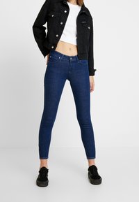 Jeans ajustados de color azul oscuro con una cintura alta, que cuentan con bolsillos frontales y cremalleras en los tobillos, combinados con una chaqueta corta negra y zapatillas negras.