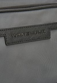 Bolsa de nailon gris con una superficie texturizada, que presenta una etiqueta de piel negra grabada con "TOMMY HILFIGER" en letras mayúsculas.