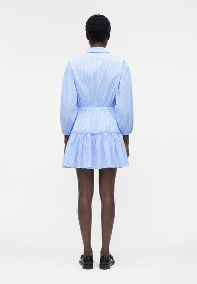 Γυναίκα που στέκεται με την πλάτη γυρισμένη, φορά ένα ανοιχτό μπλε ριγέ φόρεμα με μακριά μανίκια και βολάν στο τελείωμα, σε συνδυασμό με μαύρα loafers.