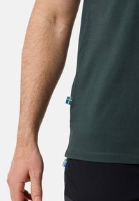 Mörkgrön bomullströja med korta ärmar, med en liten svensk flagga vid fållen, slät textur och avslappnad passform.