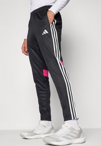 Zwarte sportieve broek met drie witte strepen aan de zijkanten en een roze accent. Gemaakt van een soepel materiaal met een elastische tailleband en zakken.