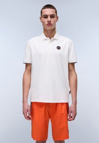 Hvid bomulds polo shirt med et brandet mærke på brystet. Sammen med lys orange shorts, der viser et afslappet, sporty look.