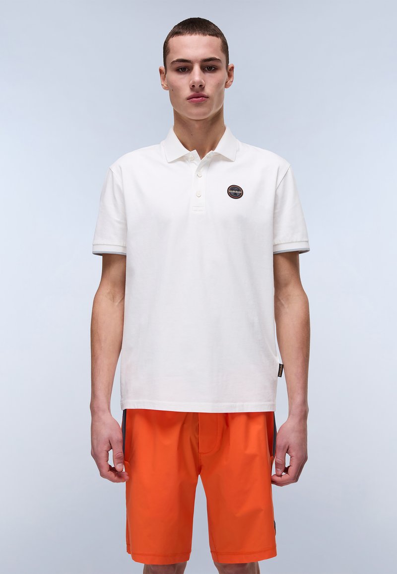 Hvid bomulds polo shirt med et brandet mærke på brystet. Sammen med lys orange shorts, der viser et afslappet, sporty look.