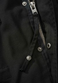 Černá látková bunda s stříbrným zipem, kovovými patentkami s logem a texturovaným prošíváním kolem zipu.