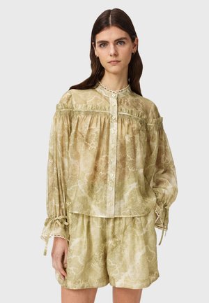 Femme portant un chemisier beige à manches longues avec boutons, motifs et détails à volants, et un short assorti, debout devant un fond uni.