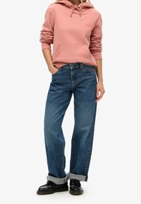 Sudadera rosa con cordón y bolsillo frontal, combinada con jeans azules holgados remangados en los puños y zapatos negros. Conjunto de vestimenta casual.