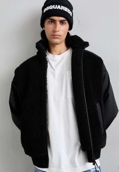 Schwarze Fleece-Weste mit Reißverschluss, die eine strukturierte Oberfläche und schwarze Stoffärmel aufweist. Über einem weißen T-Shirt getragen. Schwarze Mütze auf dem Kopf.